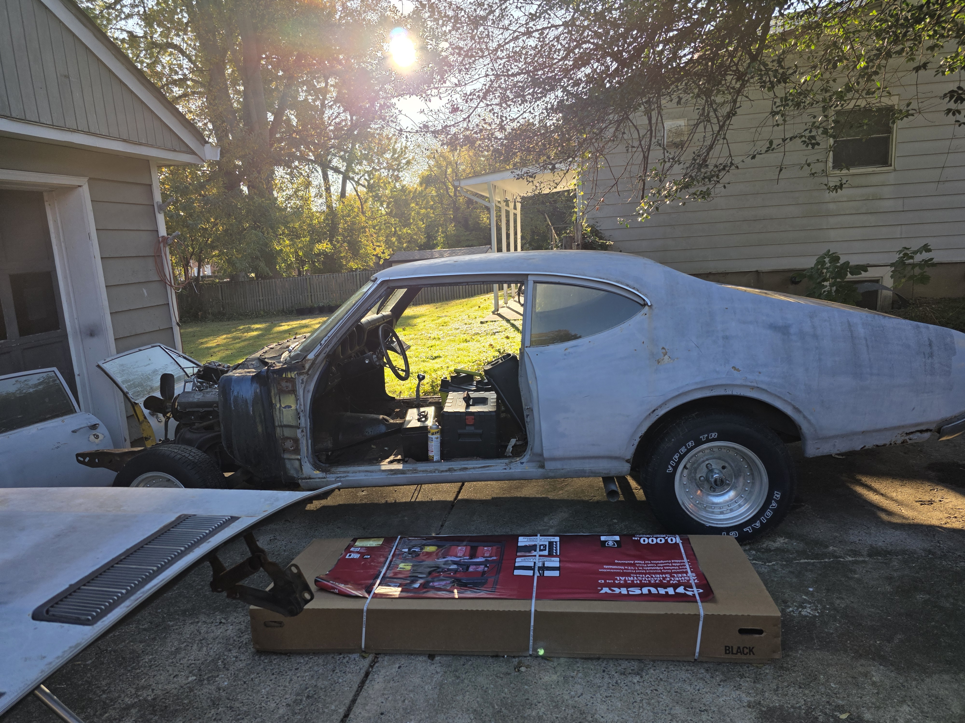 1969 Oldsmobile Cutlass S in gray primer during teardown, driver side view with the front clip removed, door off, and interior stripped, sitting in the driveway on slot mag wheels