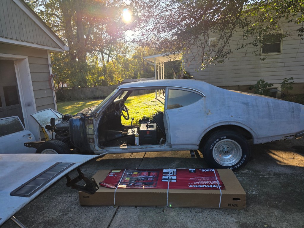 1969 Oldsmobile Cutlass S in gray primer during teardown, driver side view with the front clip removed, door off, and interior stripped, sitting in the driveway on slot mag wheels