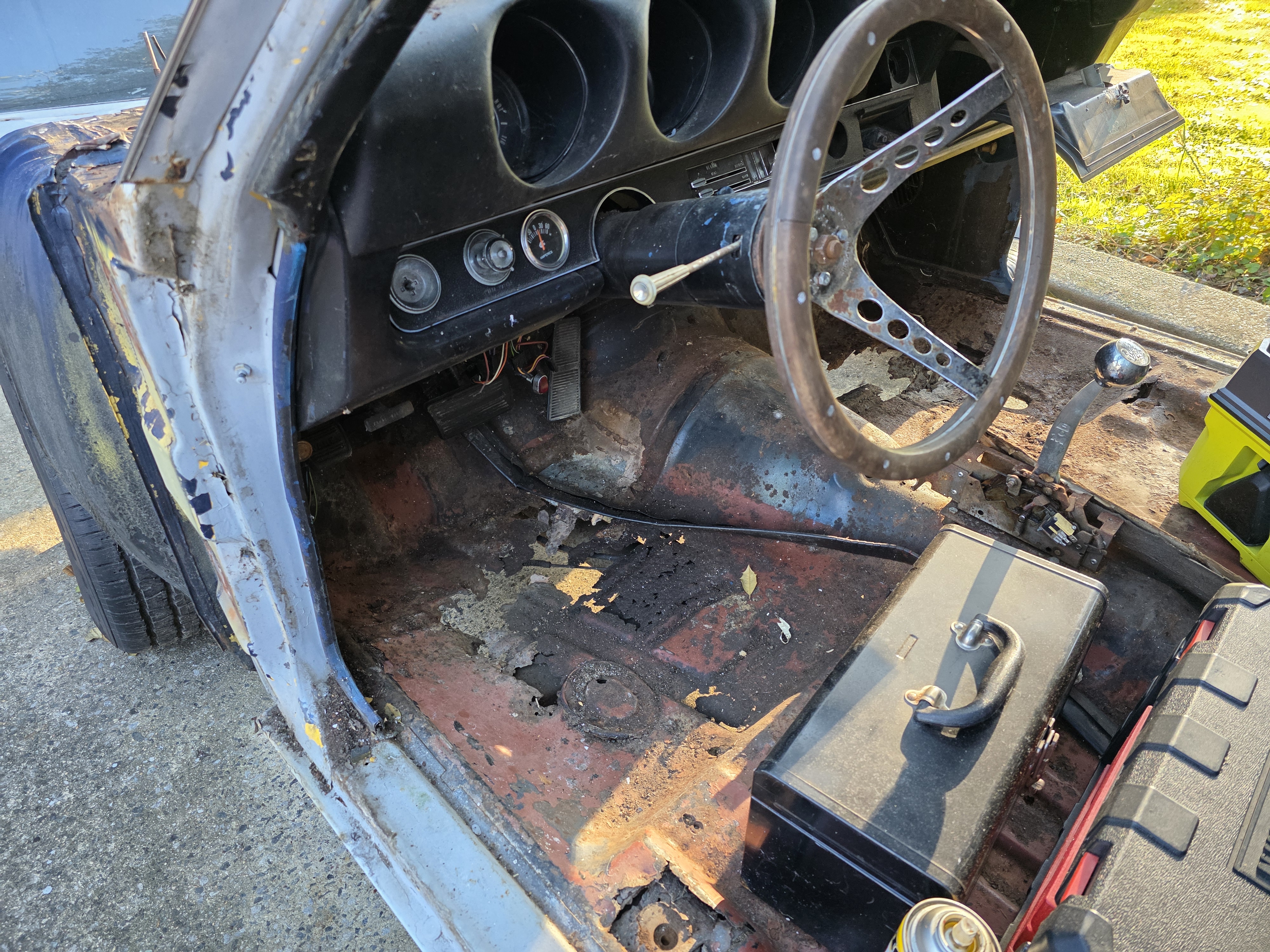 Gutted interior of a 1969 Cutlass S showing rust through holes in the driver floor pan, wood rim steering wheel and original dash still in place. Caption: The floor stopped pretending. Driver pan and passenger front half are coming out.